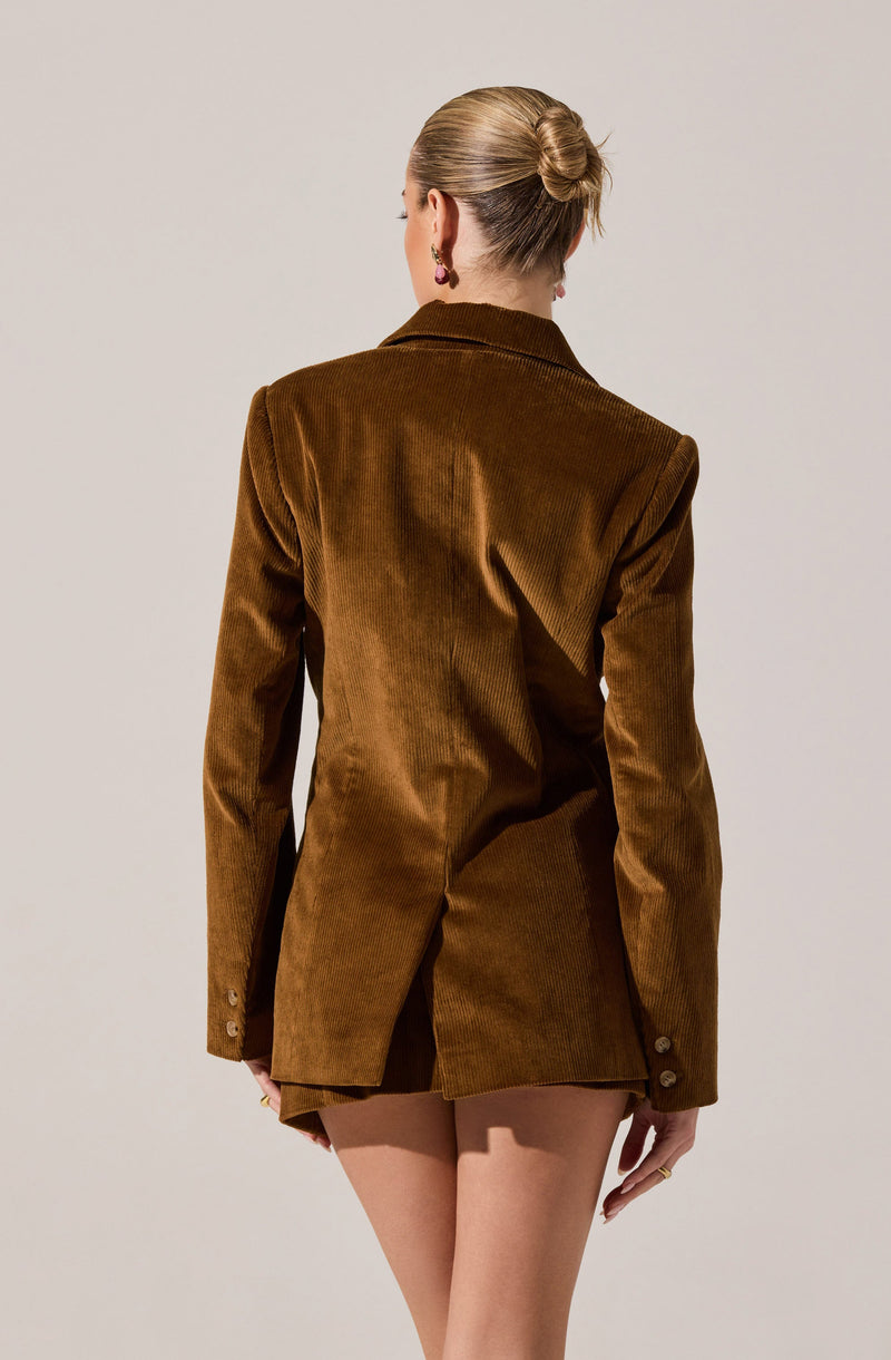 A woman is wearing a brown velvet blazer with a notched lapel collar. The blazer has long sleeves with button details at the cuffs, and it is tailored with a fitted silhouette. The back of the blazer is slightly longer, covering part of her upper thighs. She has her hair styled in a neat bun and is accessorized with earrings.