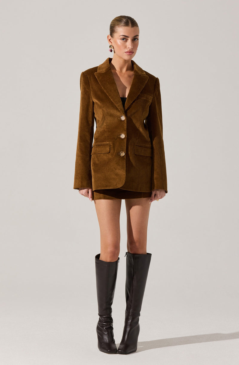 A woman is wearing a tailored brown velvet blazer with a notched lapel collar and three large buttons down the front. She is also wearing thigh-high black leather boots with a pointed toe and high heels.