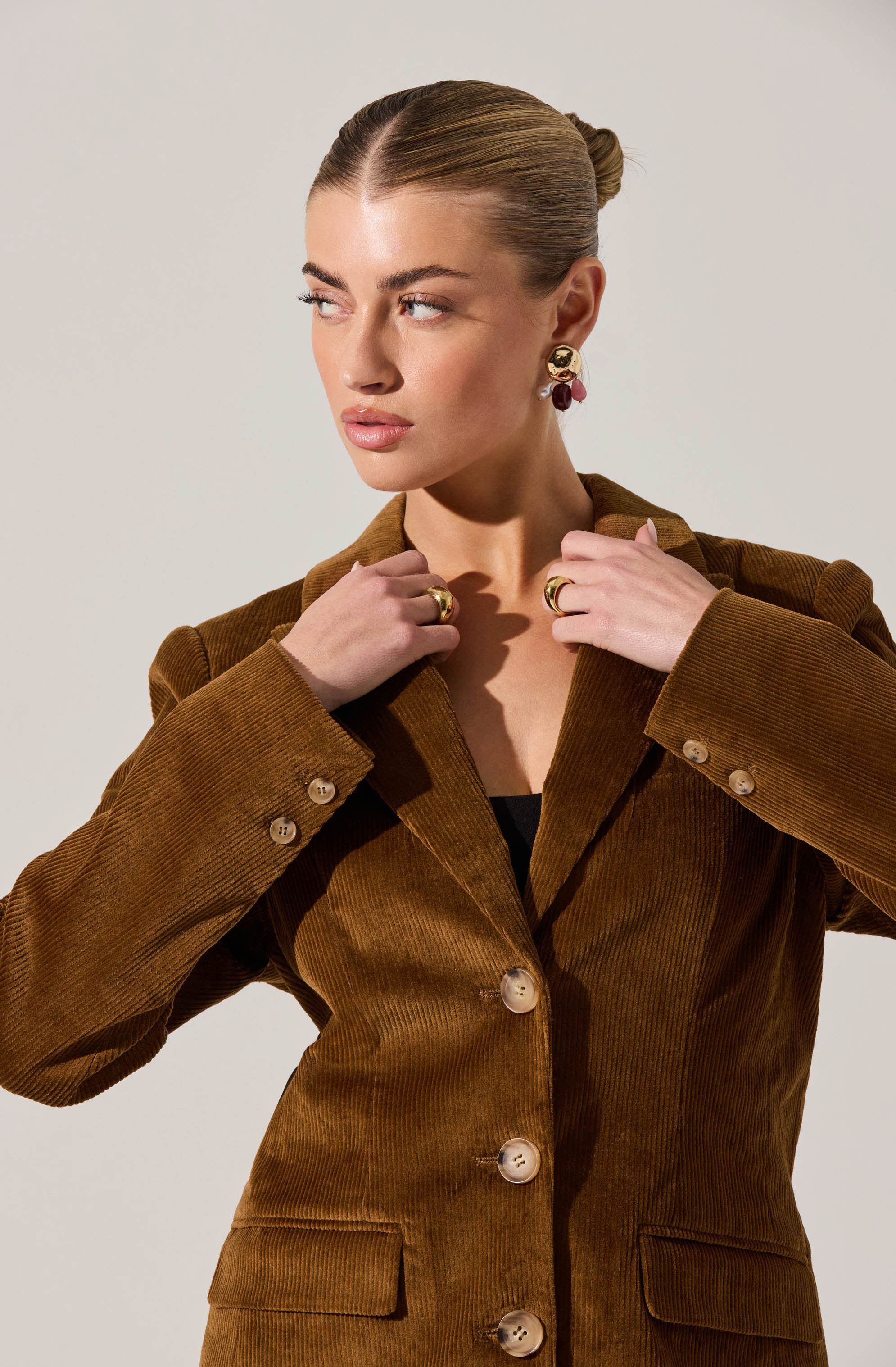 A woman is wearing a brown corduroy blazer with large buttons down the front and on the pockets. The blazer has a tailored fit with wide lapels. She is accessorized with bold earrings and rings. Her hair is styled in a sleek, low bun.