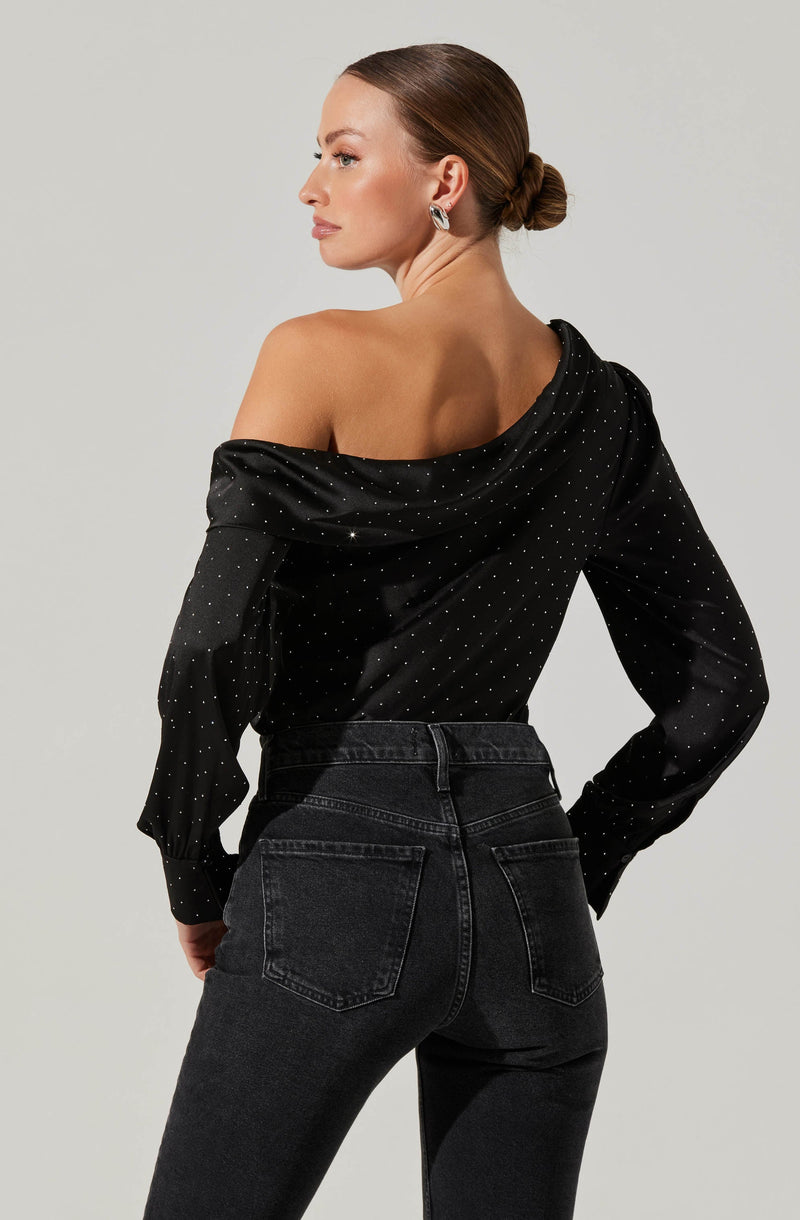A woman is wearing an off-the-shoulder black satin blouse with small white polka dots and long sleeves. She is paired with high-waisted black jeans.