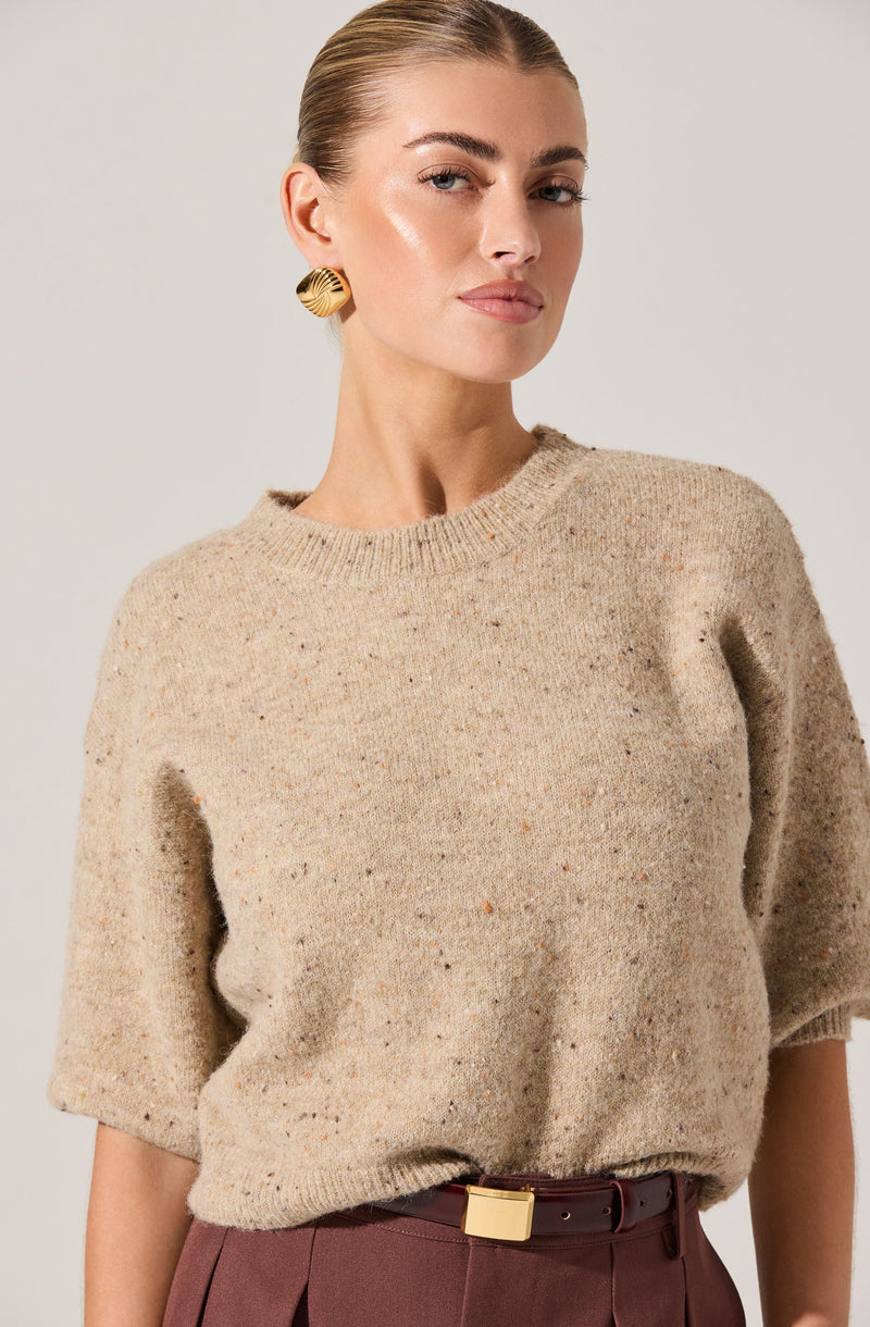 A woman is wearing a beige, fuzzy, short-sleeved sweater with a round neckline. She is also dressed in high-waisted, dark red pants with a wide belt featuring a square gold buckle. She has gold, textured, round earrings.