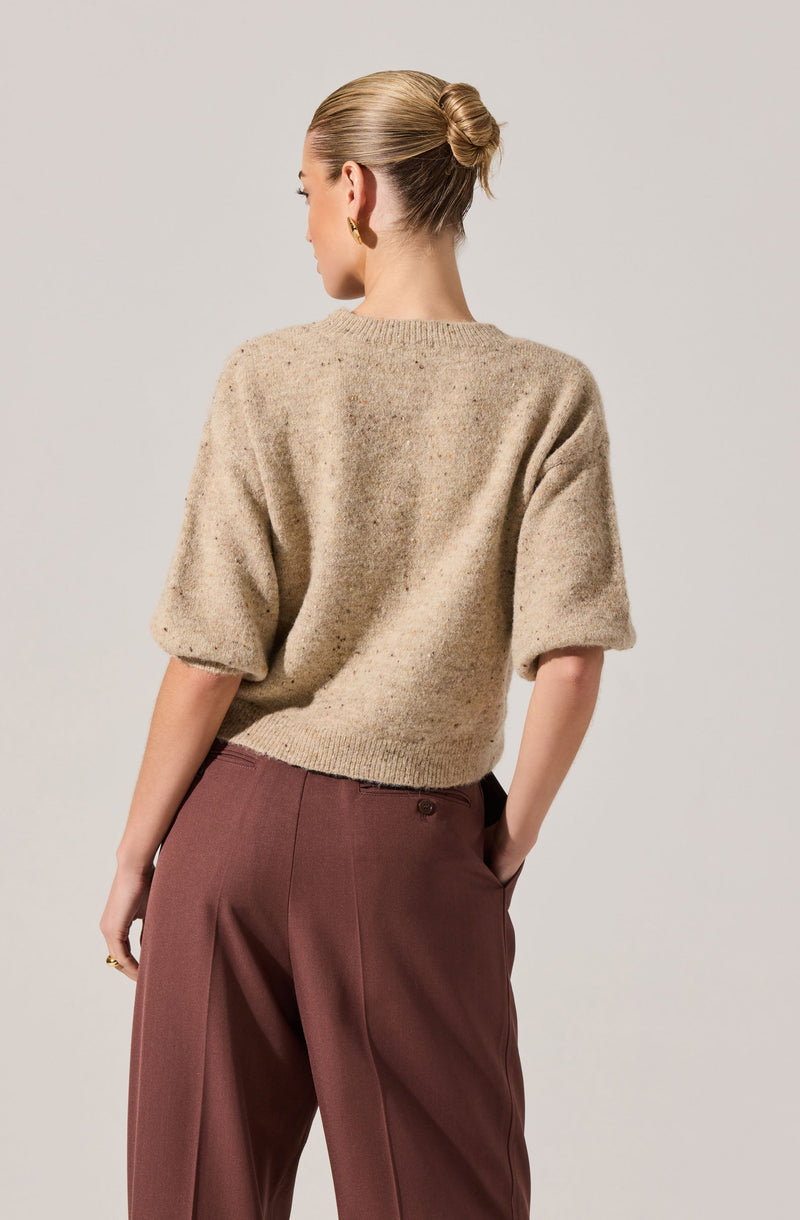 A woman is wearing a beige, textured, short-sleeved knit sweater with a round neckline. She pairs it with high-waisted, loose-fitting, brown trousers. Her hair is styled in a neat bun, and she is accessorized with gold hoop earrings.