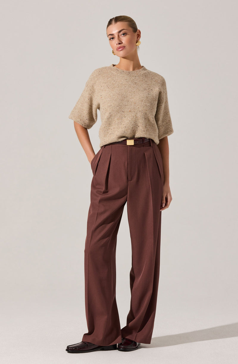 A woman is wearing a loose, beige, textured short-sleeve sweater with a round neckline, tucked into high-waisted, wide-leg brown trousers with pleats at the waist. She has a small belt with a rectangular gold buckle, and she is wearing dark brown shoes featuring a slight heel. She also has yellow earrings and is standing against a plain light background.