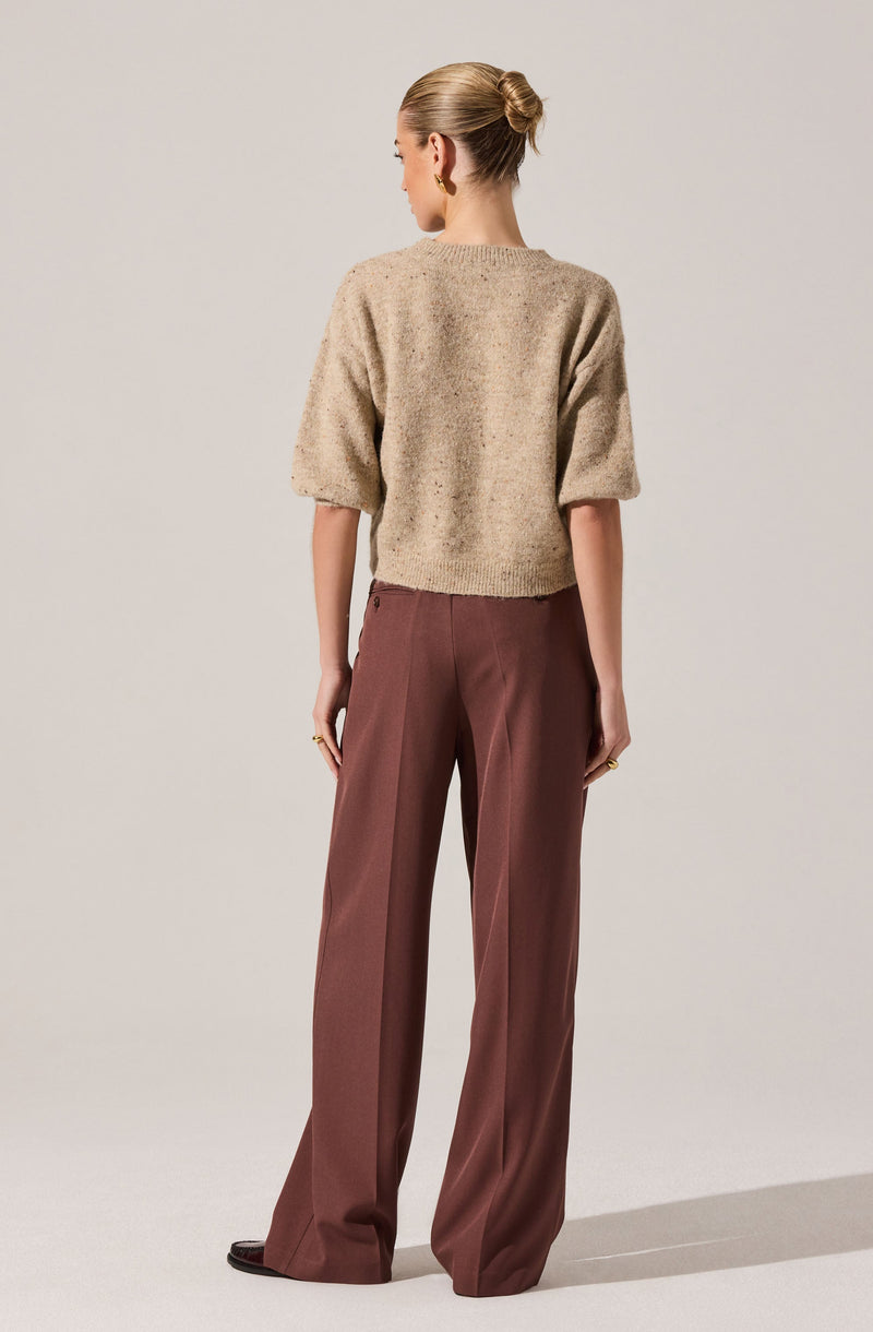 A woman is wearing a beige, loose-knit, short-sleeve sweater with a slightly cropped fit. She has paired it with high-waisted, wide-leg, brown trousers that have a tailored appearance and a subtle pleat down the front.