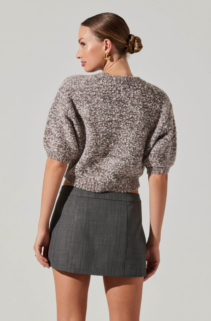A woman is wearing a fuzzy, textured, cropped sweater with puffed short sleeves in shades of gray and beige. She pairs it with a high-waisted, fitted gray plaid mini skirt. She has her hair styled in a low bun and is accessorizing with gold hoop earrings.