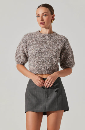 A woman is wearing a cozy, textured knit sweater in shades of brown and beige with a slightly cropped fit and puffed short sleeves. She pairs it with a high-waisted, dark gray plaid mini skirt.