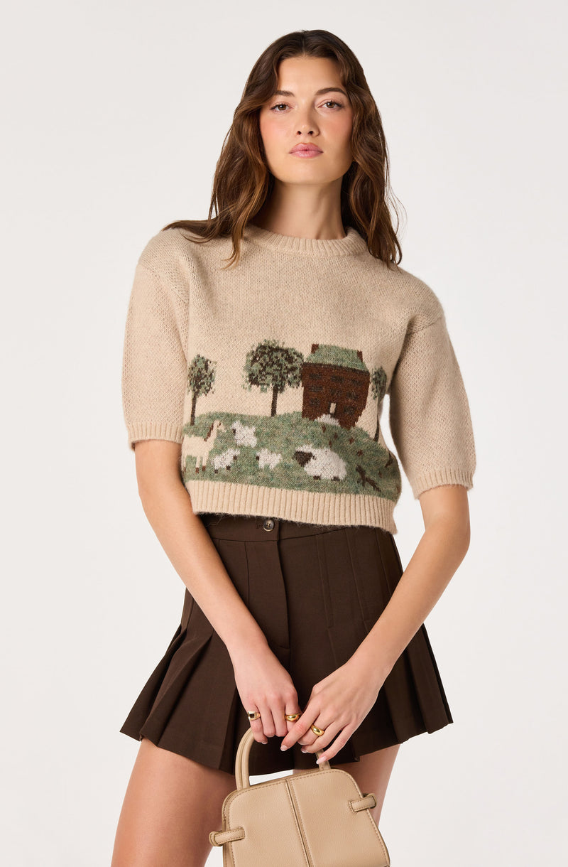Woman wearing a beige sweater with a landscape pattern and a dark brown pleated skirt, holding a beige handbag.