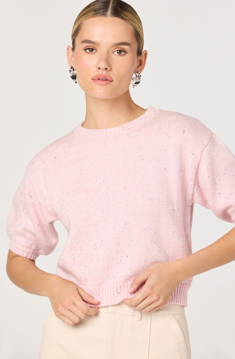 A young woman wearing a light pink knitted sweater with small multicolored specks throughout. The sweater has a round neckline and short, slightly puffed sleeves. She pairs it with beige high-waisted pants and has her blonde hair slicked back. She is also wearing large, black and white patterned earrings.