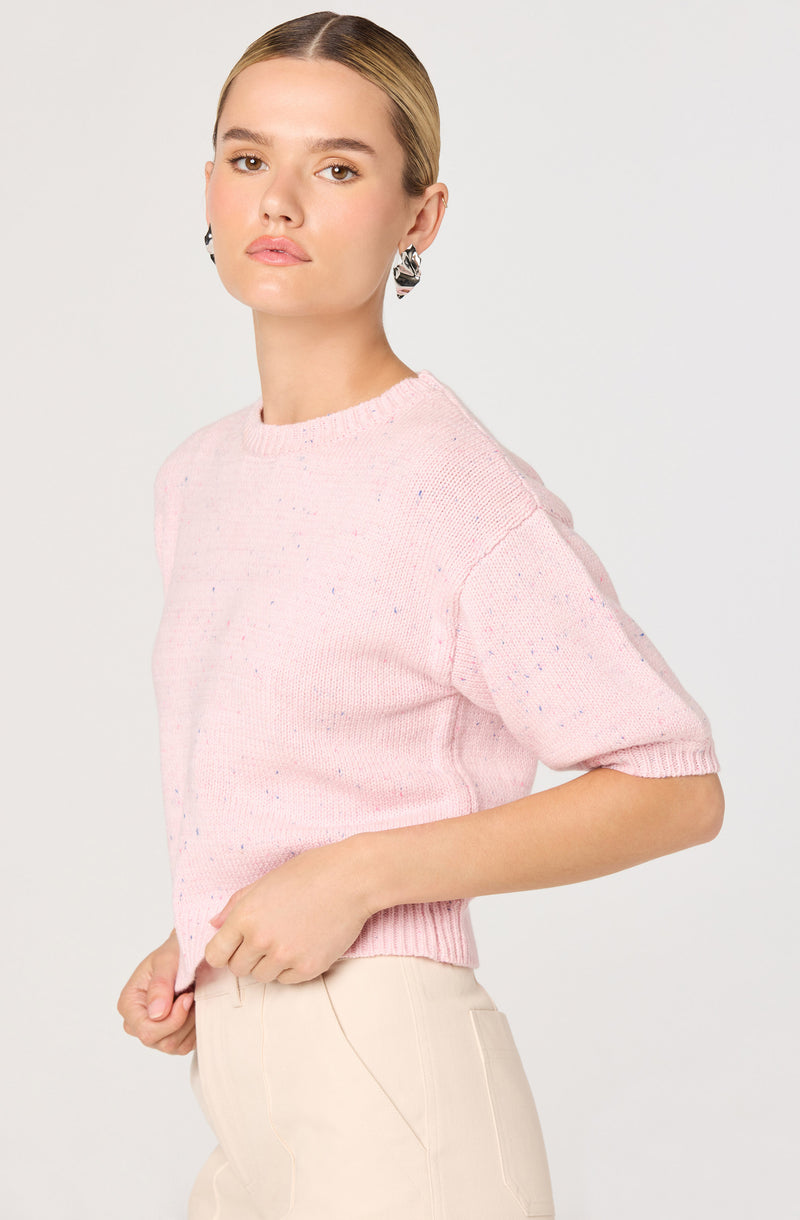 A woman is wearing a light pink, short-sleeved knit sweater with specks of multicolored threads throughout. The sweater has a crew neckline and a ribbed hem. She is also dressed in high-waisted beige pants with visible seam detailing on the front. She has short, slicked-back hair and is accessorized with large, black and white patterned earrings.