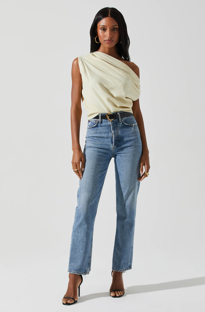 A woman is wearing a sleeveless, light yellow, draped top that exposes one shoulder. She has paired it with high-waisted, light blue jeans and a black belt with a gold circular buckle. She is also wearing black high-heeled sandals and accessorized with gold hoop earrings and rings.