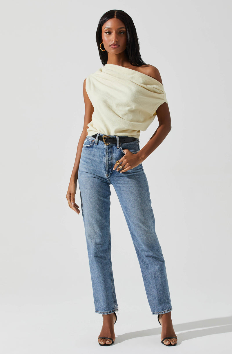A woman is wearing a cream-colored, off-the-shoulder top with a draped, loose fit. She pairs it with high-waisted, light-wash jeans and a black belt with a gold buckle. She accessorizes with gold hoop earrings and large rings. She is also wearing black high-heeled sandals.