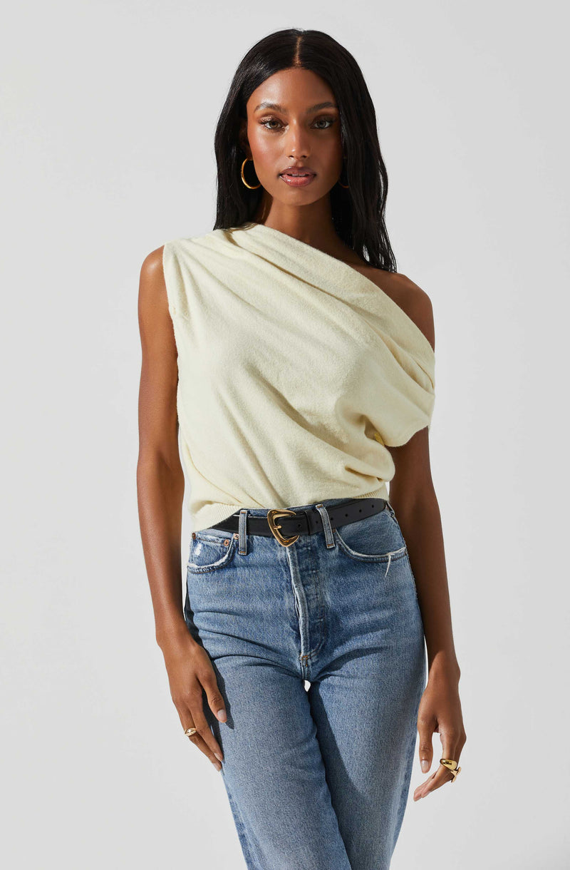 A woman wearing a cream-colored, one-shoulder, draped top paired with high-waisted, faded blue jeans secured with a black belt that has a gold buckle.