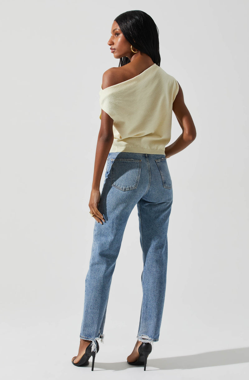 A woman is wearing a sleeveless, off-the-shoulder, light-colored knit top paired with high-waisted, slightly faded blue jeans with frayed hems.
