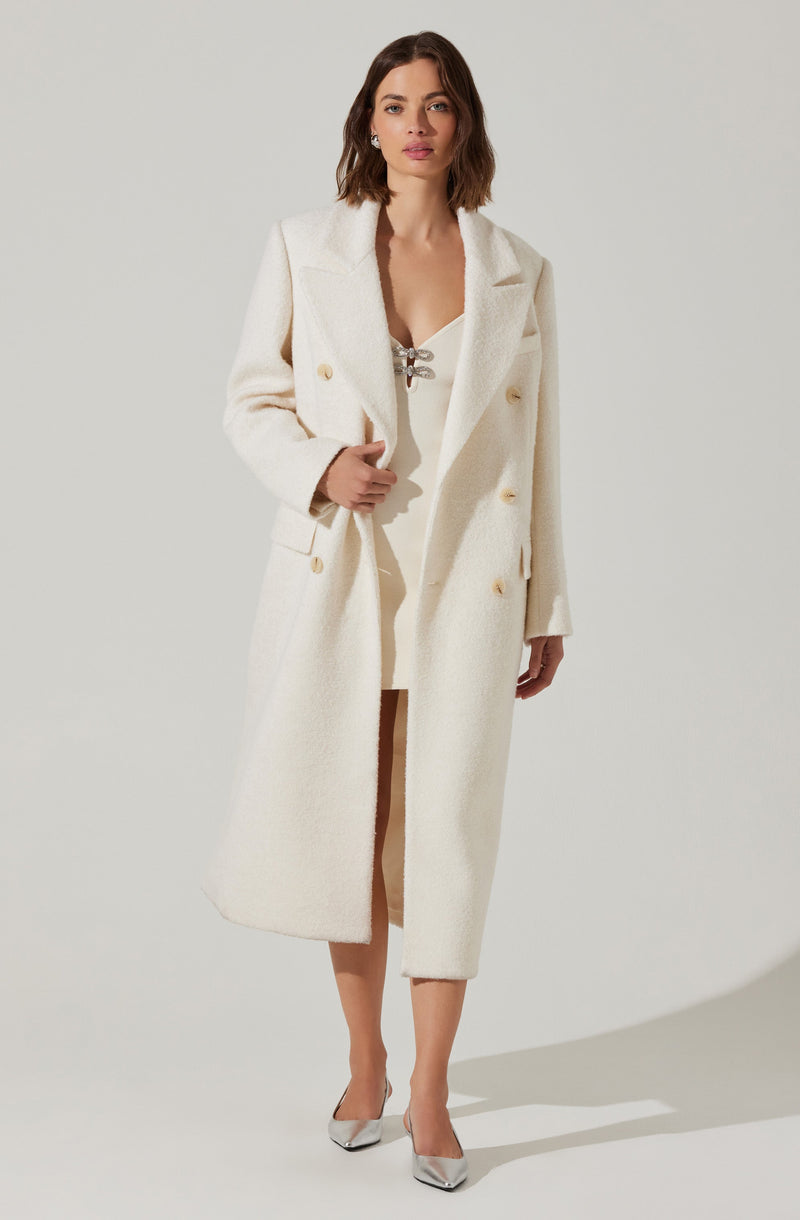 A woman is wearing a long, white, double-breasted coat with gold buttons, featuring wide lapels and a textured fabric. She has on a white dress underneath with a lace-up detail at the neckline. She is also wearing white high-heeled shoes.