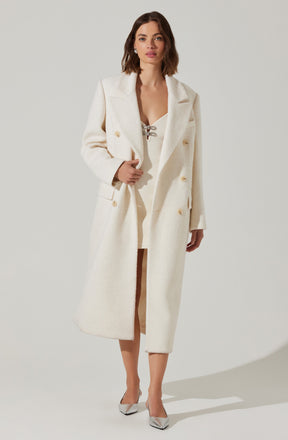 A woman is wearing a long, white, double-breasted coat with gold buttons, featuring wide lapels and a textured fabric. She has on a white dress underneath with a lace-up detail at the neckline. She is also wearing white high-heeled shoes.