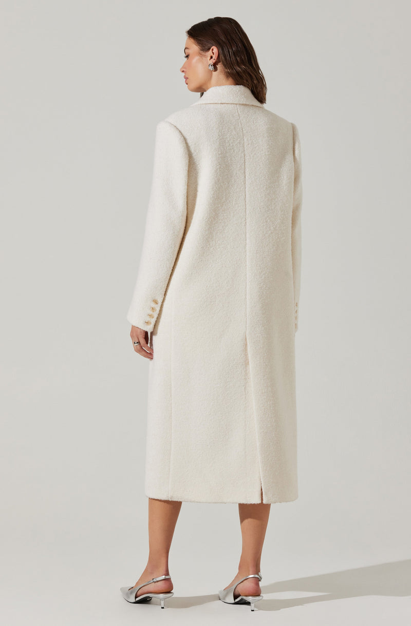 The woman is wearing a long, cream-colored coat made of a textured, possibly boucle or wool material. The coat features a notched lapel collar, and the back has a center seam with a slit at the hem. Her left sleeve has three small buttons near the cuff. She is also wearing silver, open-toed heels with ankle straps.