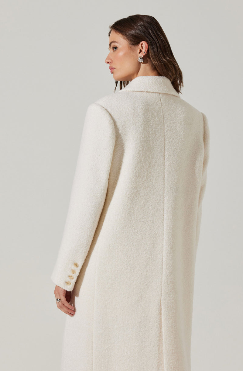 A woman is wearing a long, cream-colored, textured coat with a structured collar and shoulder pads. The coat has four buttons on each sleeve cuff.