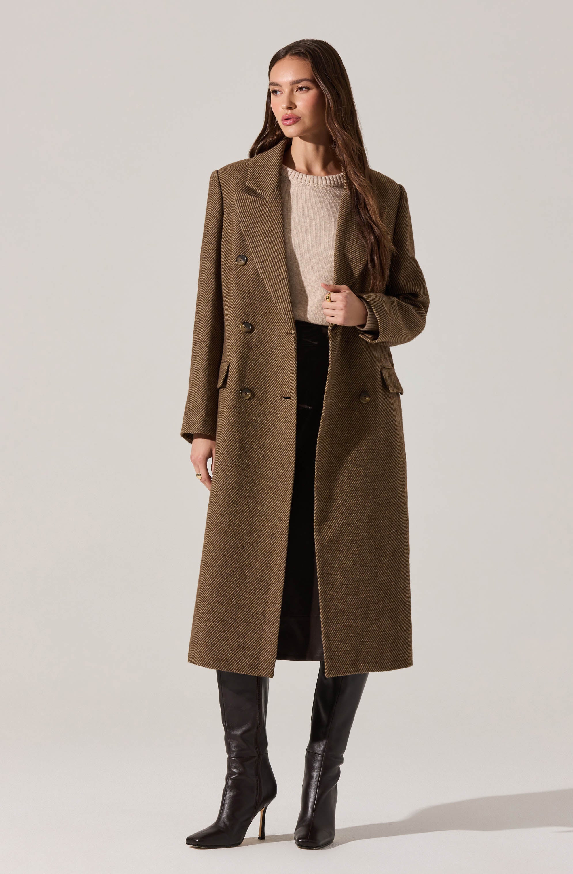 A woman is wearing a long, brown, double-breasted wool coat with a tailored fit, wide lapels, and flap pockets. Underneath, she has on a light beige knit sweater. She pairs this with black pants and black high-heeled knee-high boots.