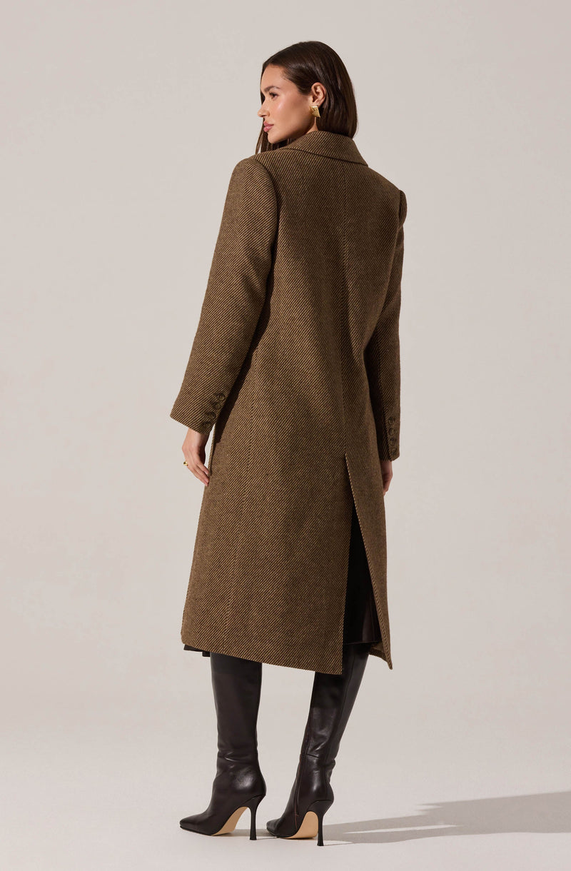 A woman is wearing a long, tailored brown coat with a subtle herringbone pattern, a classic collar, and a vent at the back. She accessorizes with gold hoop earrings and tall black leather boots with high heels.