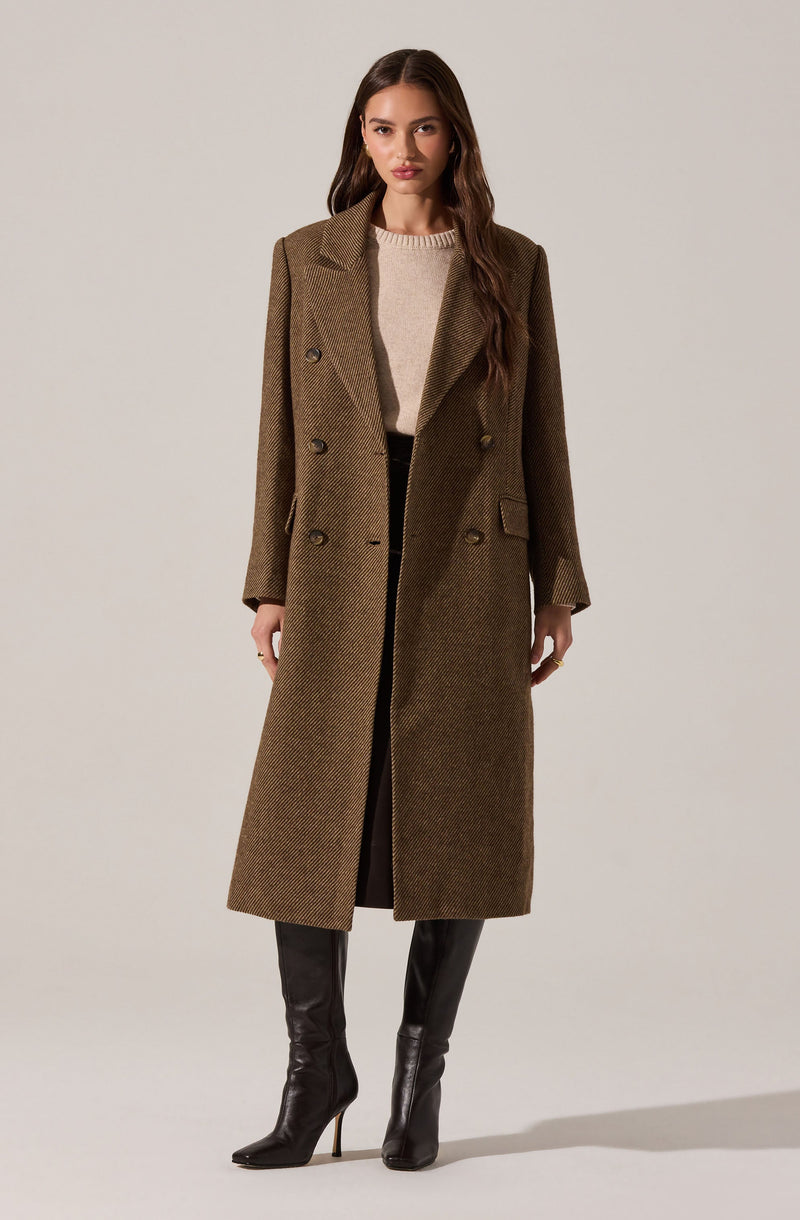 A woman is wearing a long, brown, double-breasted coat with a houndstooth pattern, featuring wide lapels and a structured fit. She has on a beige sweater underneath and black knee-high boots with high heels.