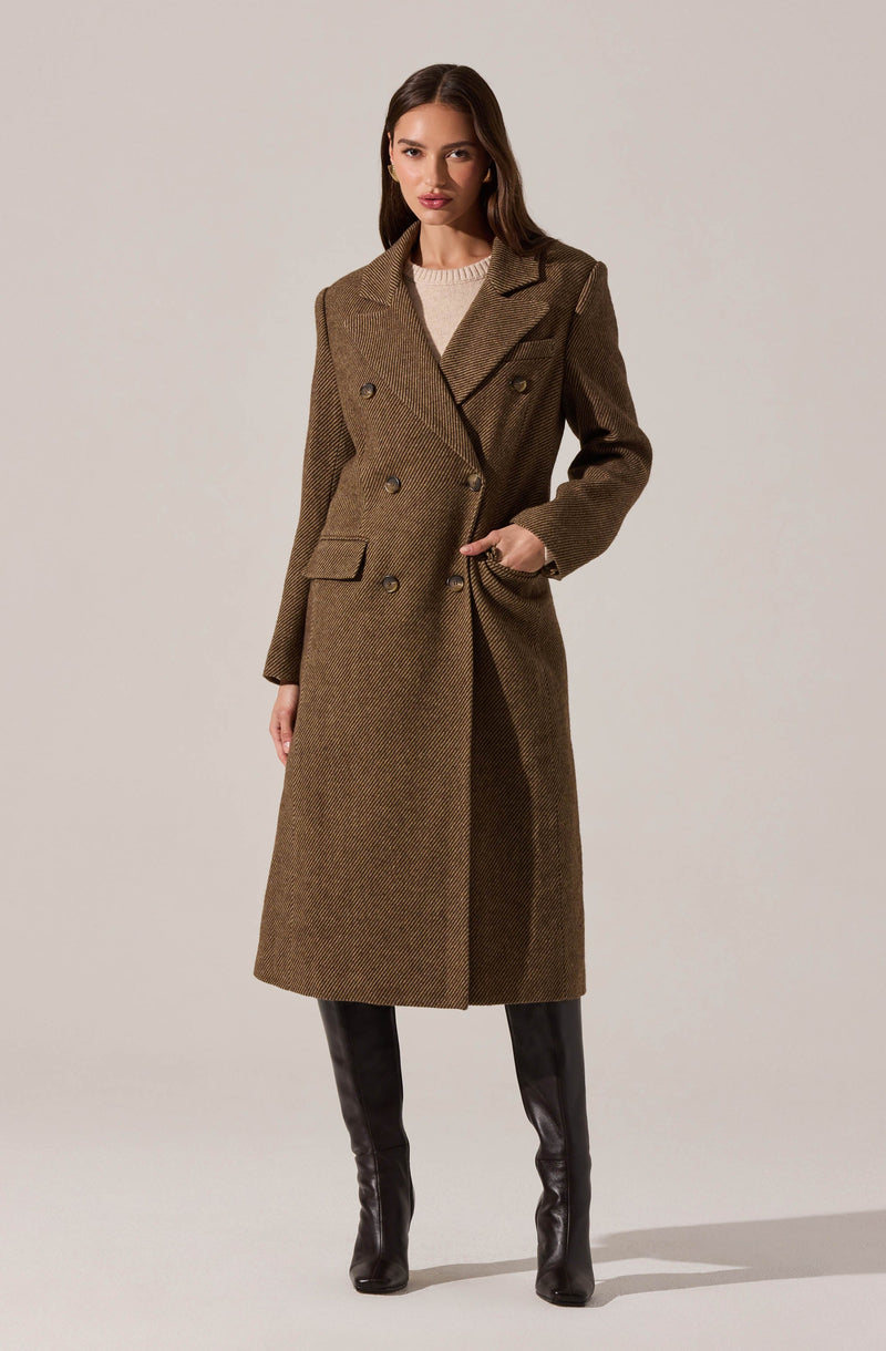 A woman is wearing a long, brown double-breasted coat with a textured, herringbone pattern. The coat features wide lapels, shoulder pads, and flap pockets. She is also dressed in black knee-high boots.