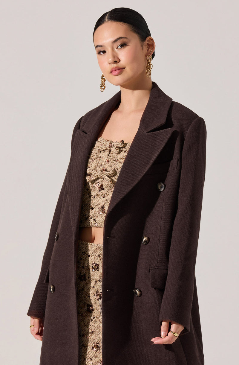A woman is wearing a dark brown, double-breasted coat with wide lapels and buttons down the front. Underneath, she has on a matching beige, textured, cropped top and skirt set with a floral pattern. She accessorizes with large gold hoop earrings.