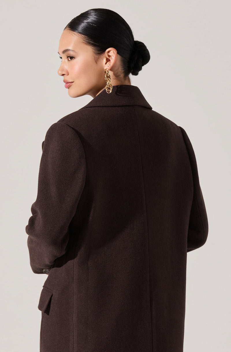A woman with dark hair styled in a low bun is wearing a dark brown, textured blazer with a notched collar and long sleeves. She has large gold hoop earrings.