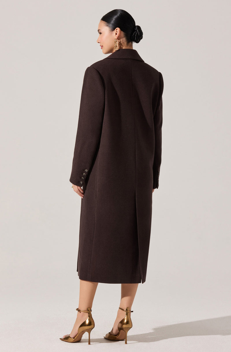 A woman wearing a long, dark brown blazer-style coat that reaches mid-calf. The coat features a tailored fit with a notched collar, long sleeves with buttons on the cuffs, and a subtle vent at the back. She is also wearing gold high-heeled sandals.
