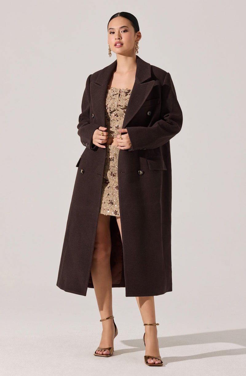 A woman is wearing a long, dark brown, double-breasted coat with wide lapels and buttons on the front. Underneath, she has on a knee-length, beige dress with a floral or tapestry pattern, and she is wearing high-heeled sandals with ankle straps. She has accessorized with large, gold dangling earrings.