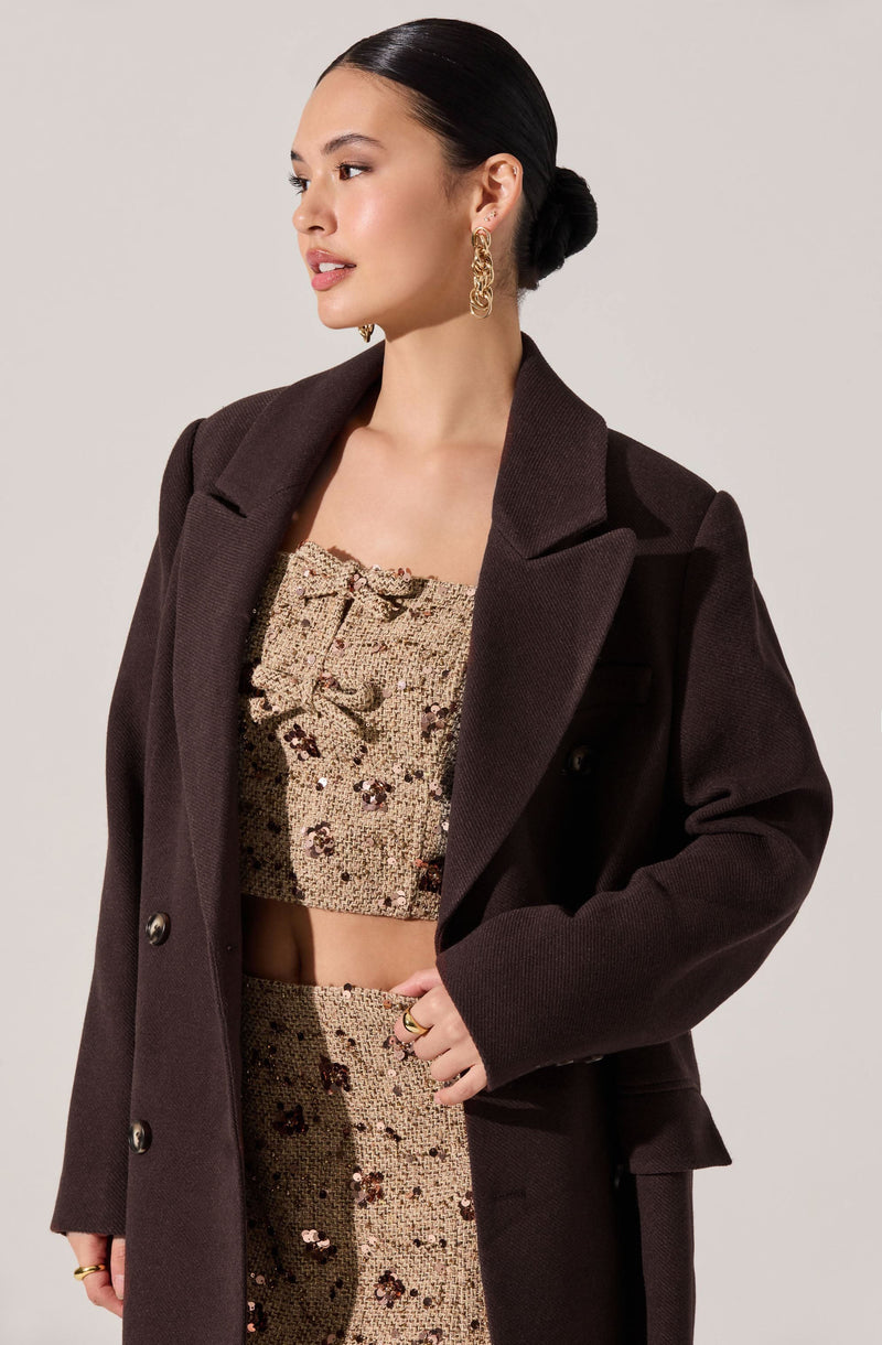 A woman is wearing a dark brown, double-breasted blazer with wide lapels and long sleeves. Underneath, she has on a sleeveless, cropped top and matching high-waisted pants made of fabric with a beige background and a floral pattern in shades of purple and brown. She has accessorized with large, gold, circular earrings and multiple rings. Her hair is sleek and pulled back into a low bun.