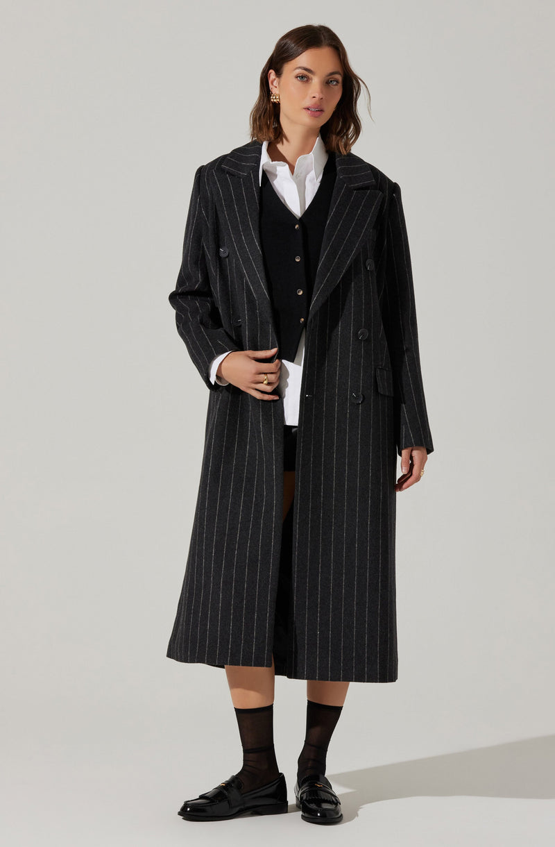 A woman is wearing a long, black pinstripe trench coat over a white collared shirt and a black cardigan. She has paired the outfit with black socks and black loafers. The coat features a classic lapel collar and double-breasted buttons.