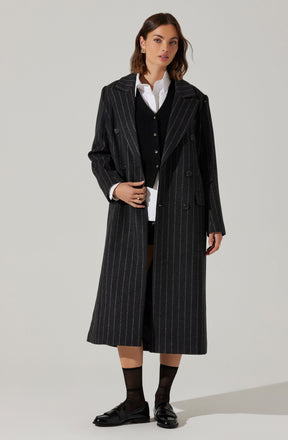 A woman is wearing a long, black pinstripe trench coat over a white collared shirt and a black cardigan. She has paired the outfit with black socks and black loafers. The coat features a classic lapel collar and double-breasted buttons.