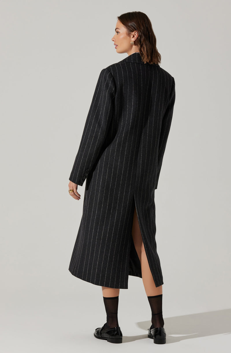 A woman is wearing a long, dark gray pinstripe coat with a slit at the back. The coat is tailored with a structured fit, featuring vertical white pinstripes, and extends to her ankles. She has dark socks and black loafers.