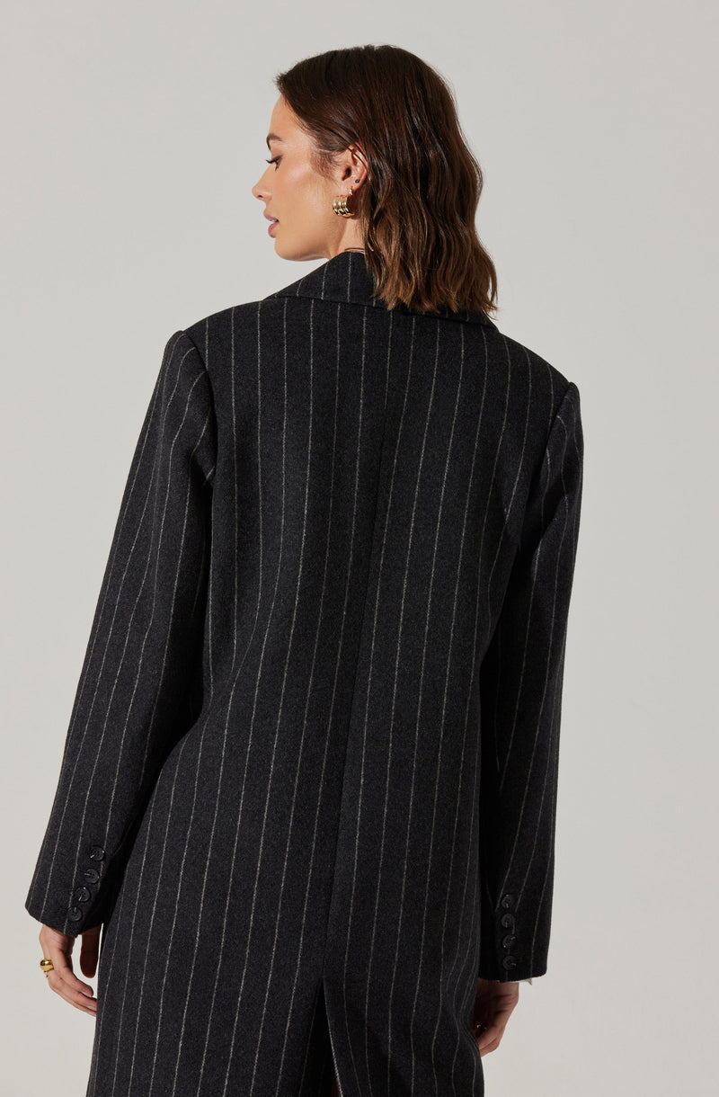 A woman is wearing a dark gray pinstripe blazer with light-colored vertical stripes. The blazer features a notched lapel collar and has buttons on the cuffs of the long sleeves. The back of the blazer extends down past her waist, with a slit at the center.