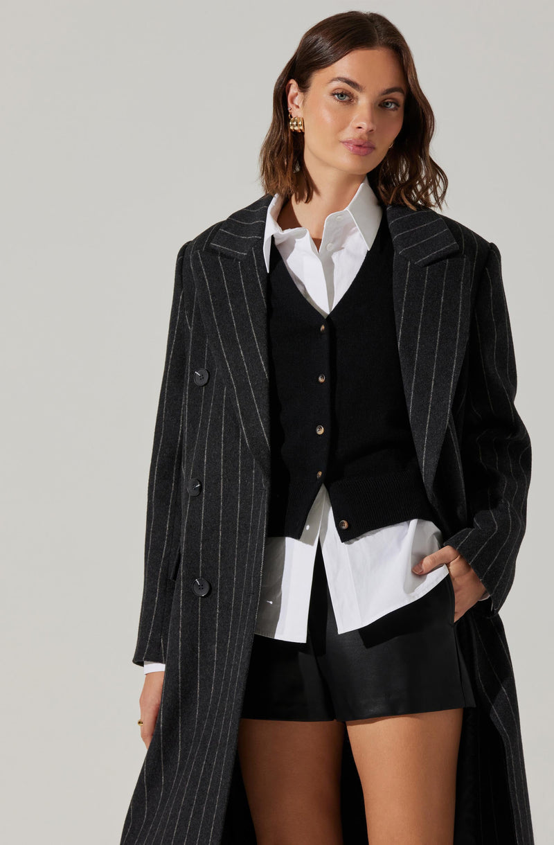 A woman is wearing a dark gray, pinstripe, double-breasted coat with wide lapels. Underneath, she has on a white collared shirt layered with a black button-up cardigan. She is also wearing black shorts. The overall look combines formal and casual elements.