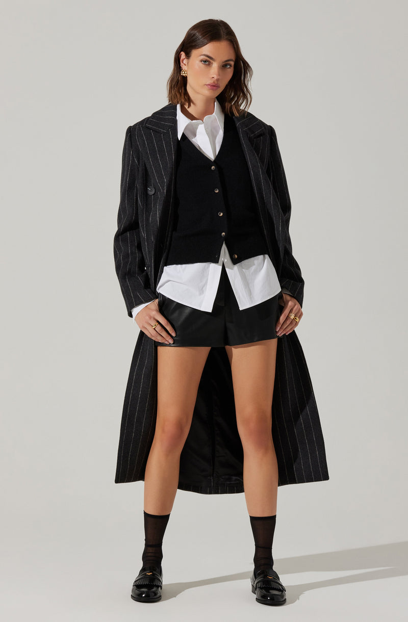 A woman is wearing a long, dark pinstripe coat over a black cardigan and a white collared shirt. She is also wearing black shorts, black socks, and black shoes.