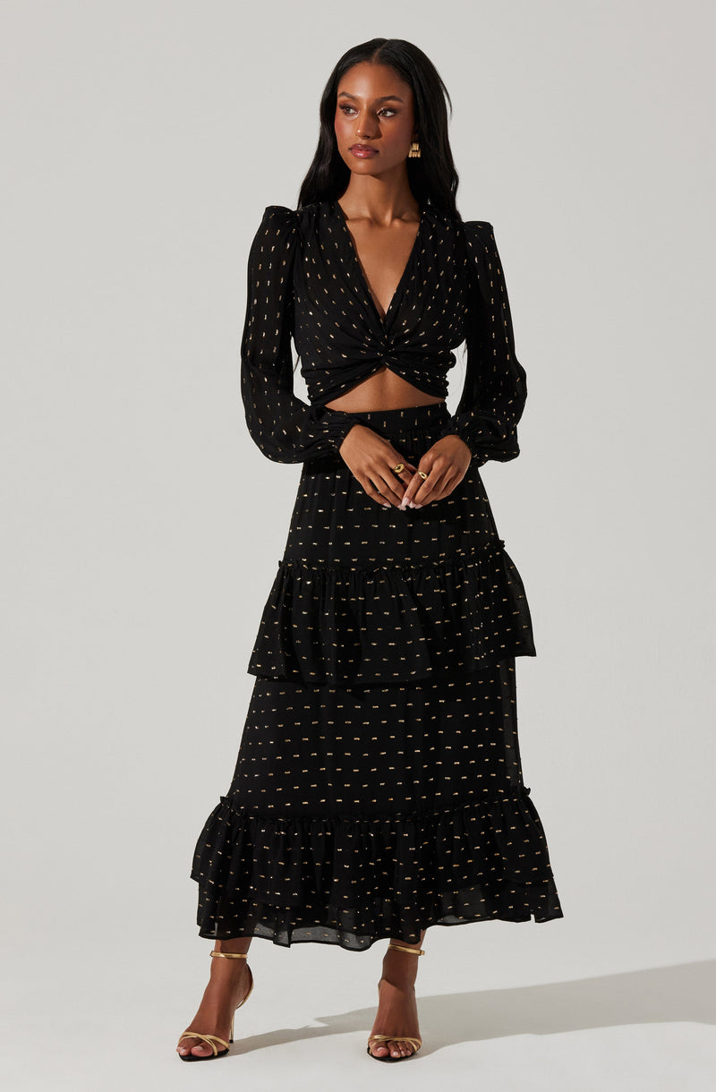 A woman is wearing a black two-piece outfit with a gold polka dot pattern. The top has long puffed sleeves, a V-neckline, and is twisted at the waist, exposing some midriff. The skirt is high-waisted, tiered with ruffles, and falls to her ankles.