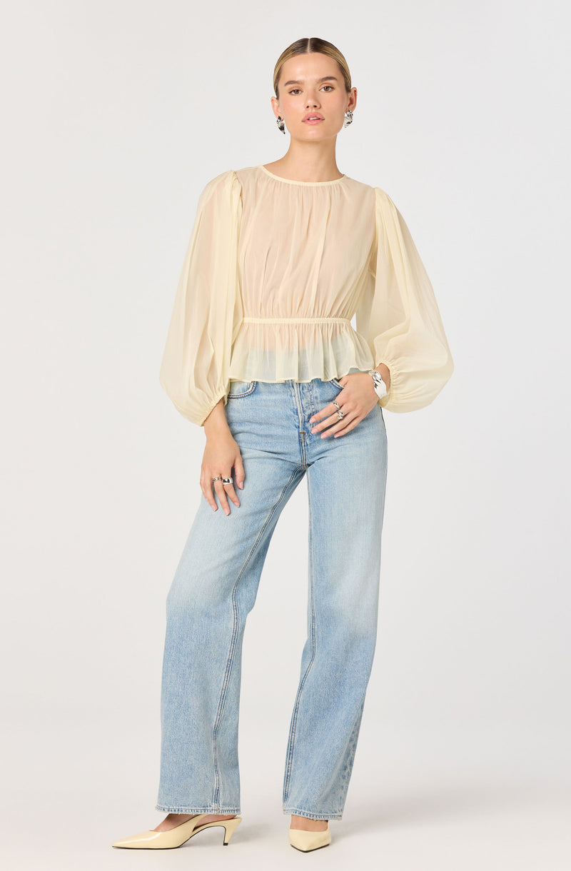 A woman is wearing a cream-colored, long-sleeved blouse with puffed, voluminous sleeves and a ruffled hem. The blouse has a gathered neckline and a slightly cinched waist. She pairs it with light-wash, high-waisted, straight-leg jeans. She completes the outfit with cream-colored pointed-toe heels and accessorizes with earrings, rings, and a watch.