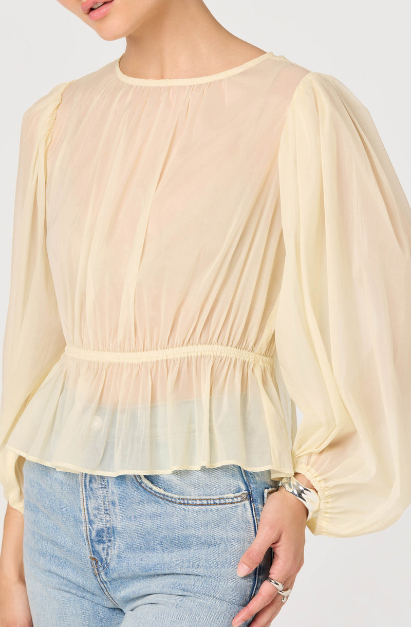 A woman is wearing a light, sheer, cream-colored blouse with long, voluminous bishop sleeves that are gathered at the wrists. The blouse features vertical pleats on the front, a rounded neckline, and a ruffled hemline that cinches at the waist. She is also wearing light blue denim jeans and a silver bracelet on her wrist.