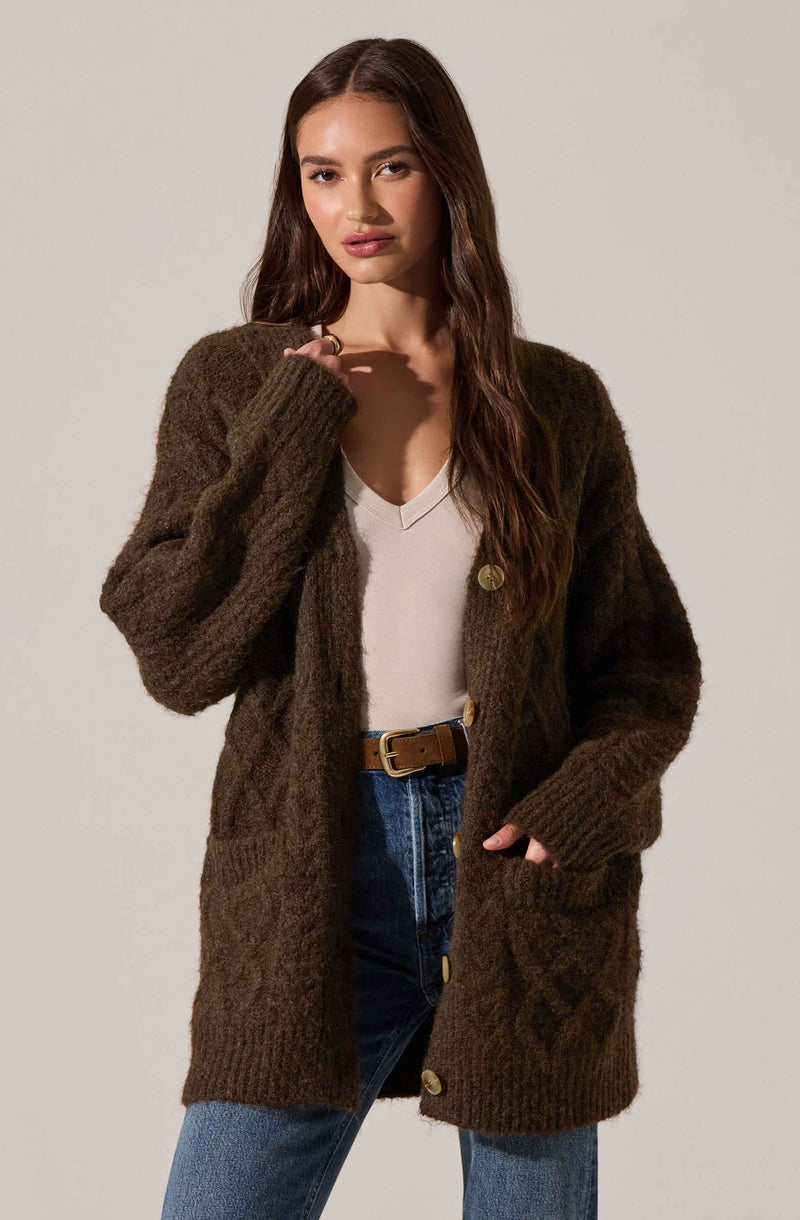 A woman is wearing a chunky, oversized brown knit cardigan with large buttons, a light beige v-neck top, and high-waisted blue jeans with a brown belt.