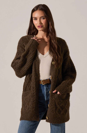 A woman is wearing a chunky, oversized brown knit cardigan with large buttons, a light beige v-neck top, and high-waisted blue jeans with a brown belt.