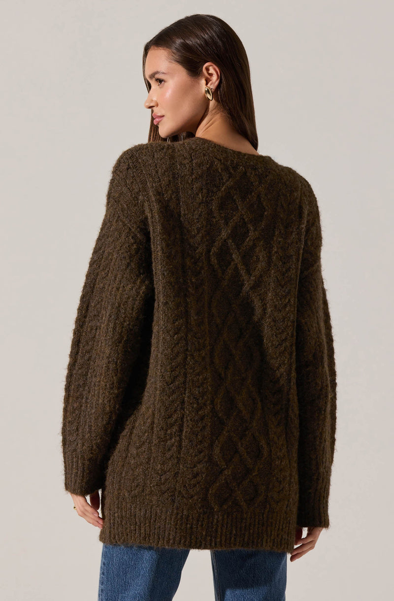 A woman is wearing a oversized, knit cardigan sweater in a dark brown color. The sweater features a textured cable knit pattern down the back and sleeves. She is also wearing blue jeans and has small hoop earrings.