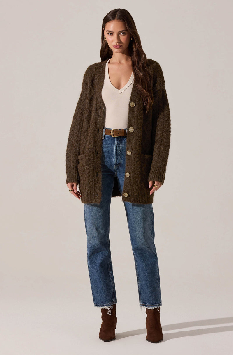A woman is wearing a long, chunky brown cardigan with large buttons, a white V-neck top, high-waisted blue jeans with a frayed hem, a brown belt, and ankle-high brown boots.