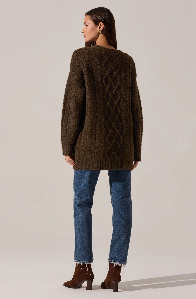 A woman is wearing a long, oversized, knitted cardigan in a dark brown color, featuring a textured cable knit pattern down the back. She pairs it with blue jeans and brown ankle boots with a chunky heel.