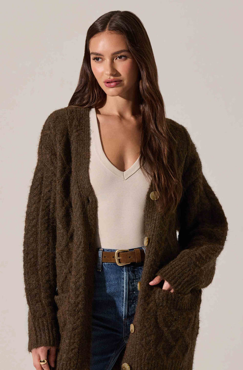 A woman is wearing a long, oversized, dark brown, knitted cardigan with large buttons down the front. Underneath, she has on a white V-neck top. She is also wearing high-waisted blue jeans with a brown belt.