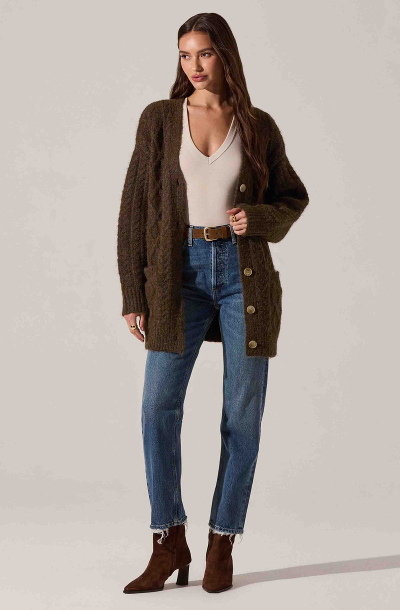 A woman is wearing a long, chunky knit cardigan in dark brown with large buttons down the front. Underneath, she has on a light-colored, V-neck top. She is also dressed in high-waisted, slightly faded blue jeans with raw hem at the ankles, secured with a brown belt. She completes her outfit with dark brown ankle boots with a high heel.