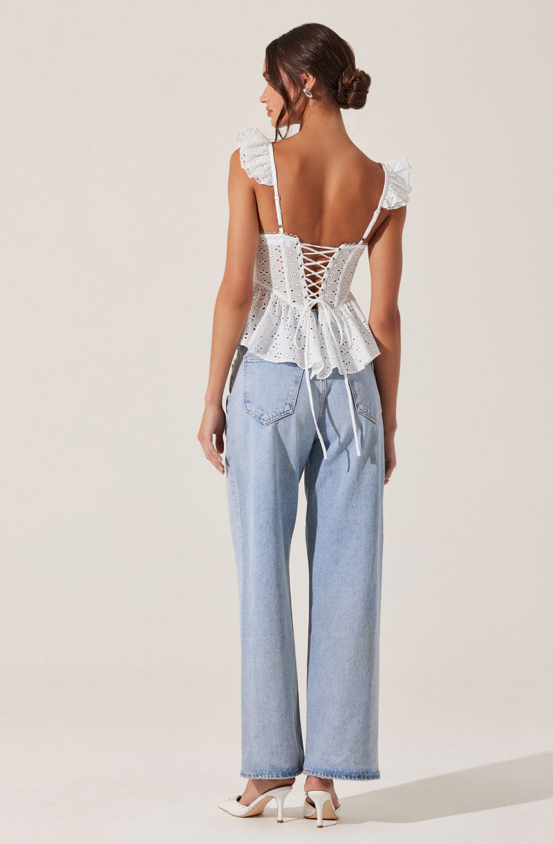 A woman is wearing a white eyelet top with ruffled shoulder straps and a fitted bodice that laces up the back with a peplum hem. She pairs it with light-wash, high-waisted, wide-leg jeans and white high-heeled shoes.