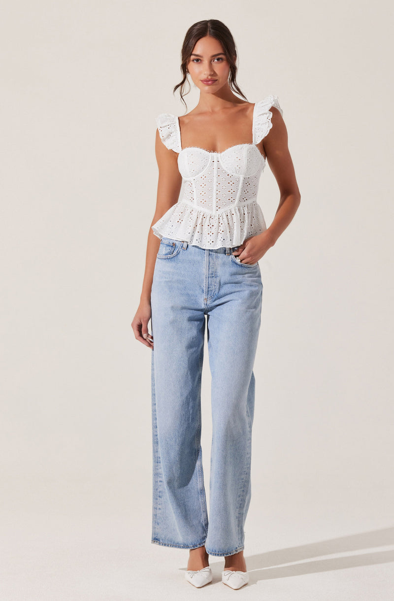 A woman is wearing a white eyelet corset top with ruffled shoulder straps and a peplum hem. She pairs it with light-wash, high-waisted wide-leg jeans and white pointed-toe shoes.