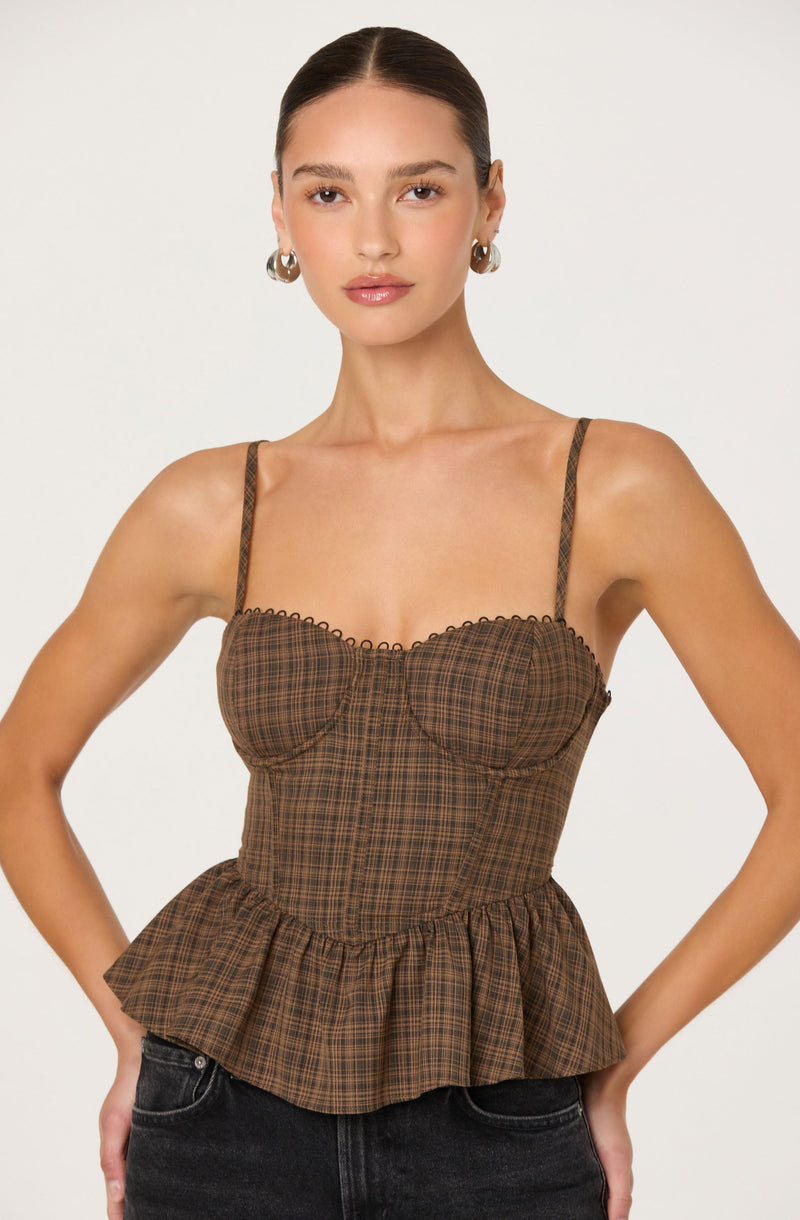 A woman is wearing a fitted brown plaid sleeveless top with thin spaghetti straps. The top features a sweetheart neckline with subtle scalloped trim and a ruffled peplum hem that cinches at the waist. She pairs it with dark-colored jeans and accessorizes with silver hoop earrings.