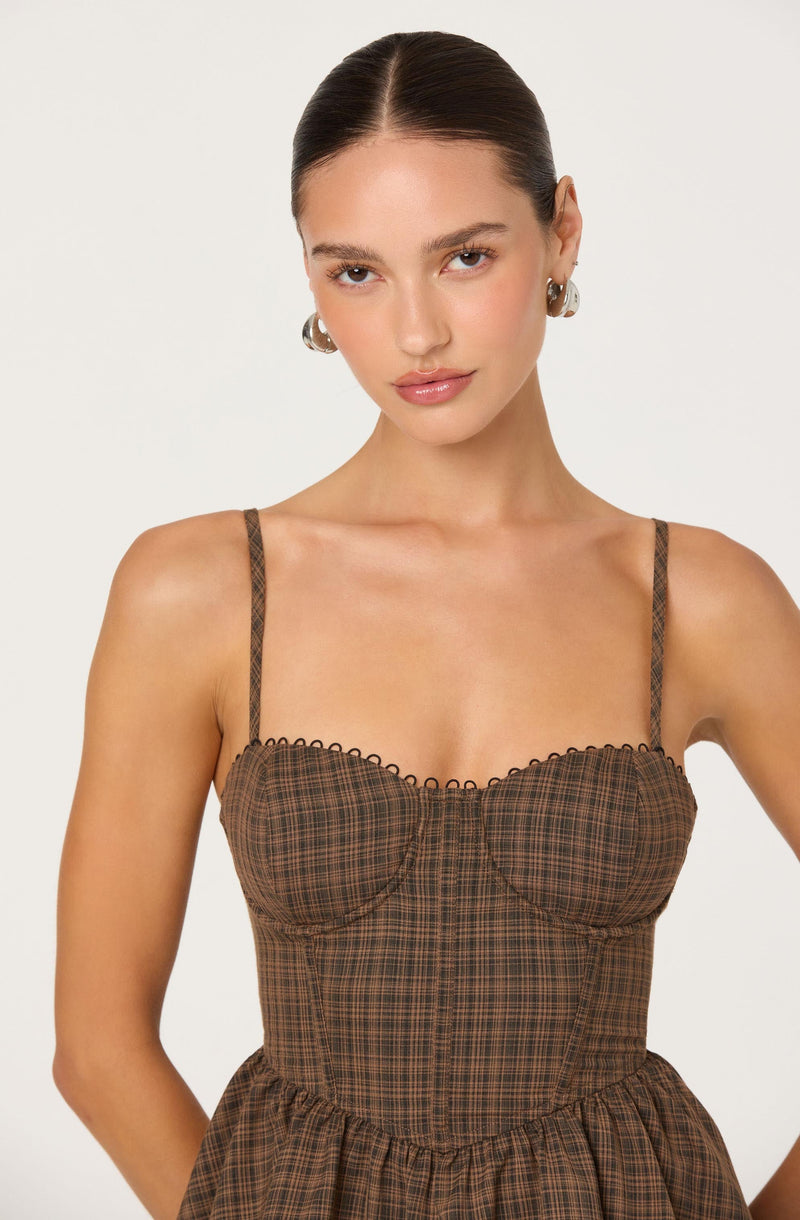 A young woman is wearing a brown plaid dress with a fitted bodice and thin, braided spaghetti straps. The dress features a scalloped trim along the top edge and a gathered, elastic waistband. The fabric has a small checkered pattern in shades of brown and black. She has her hair pulled back and is accessorized with large, silver hoop earrings.